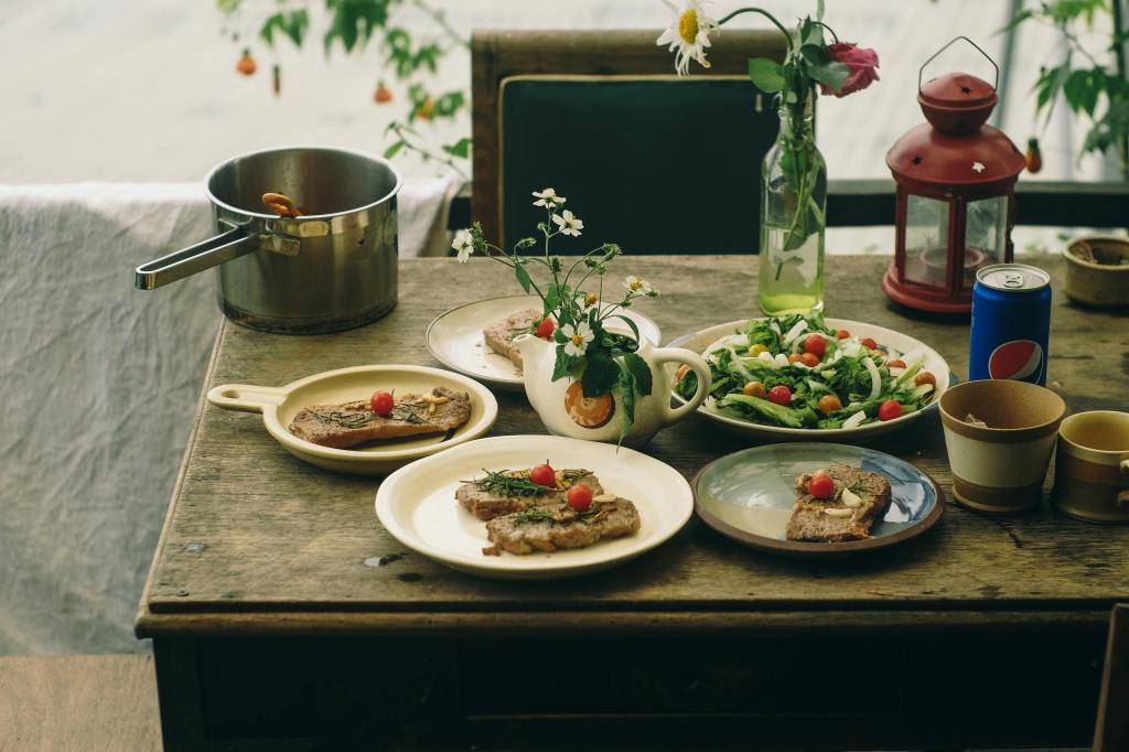 Plates of meat, vegetables, and fruit on a wooden table, with drinks and a pot nearby.