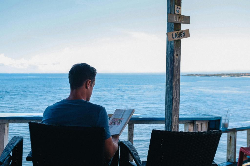 A man sitting outside reading a book next to the ocean.