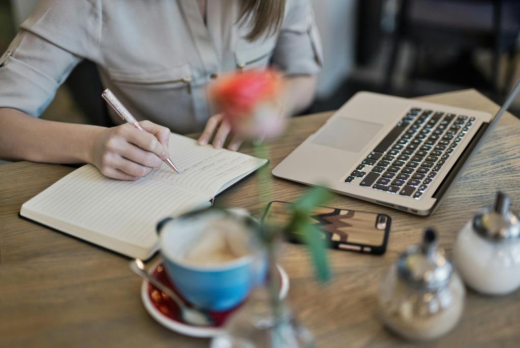 A woman sitting at a desk, writing in a journal with her laptop open and close by.