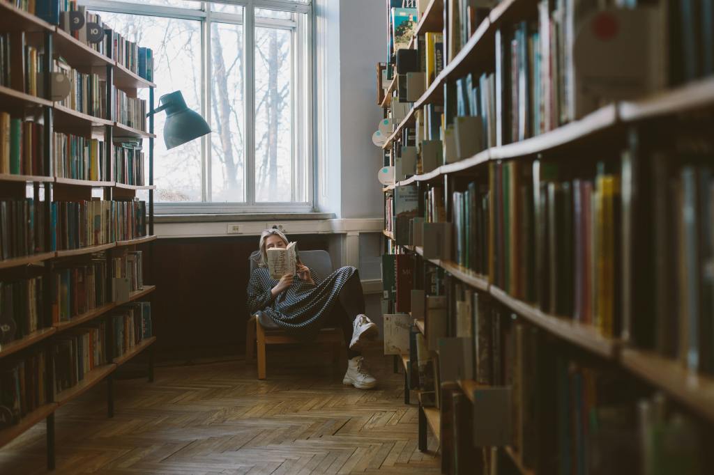 A young lady at the end of a hallway lined up with bookshelves reading.